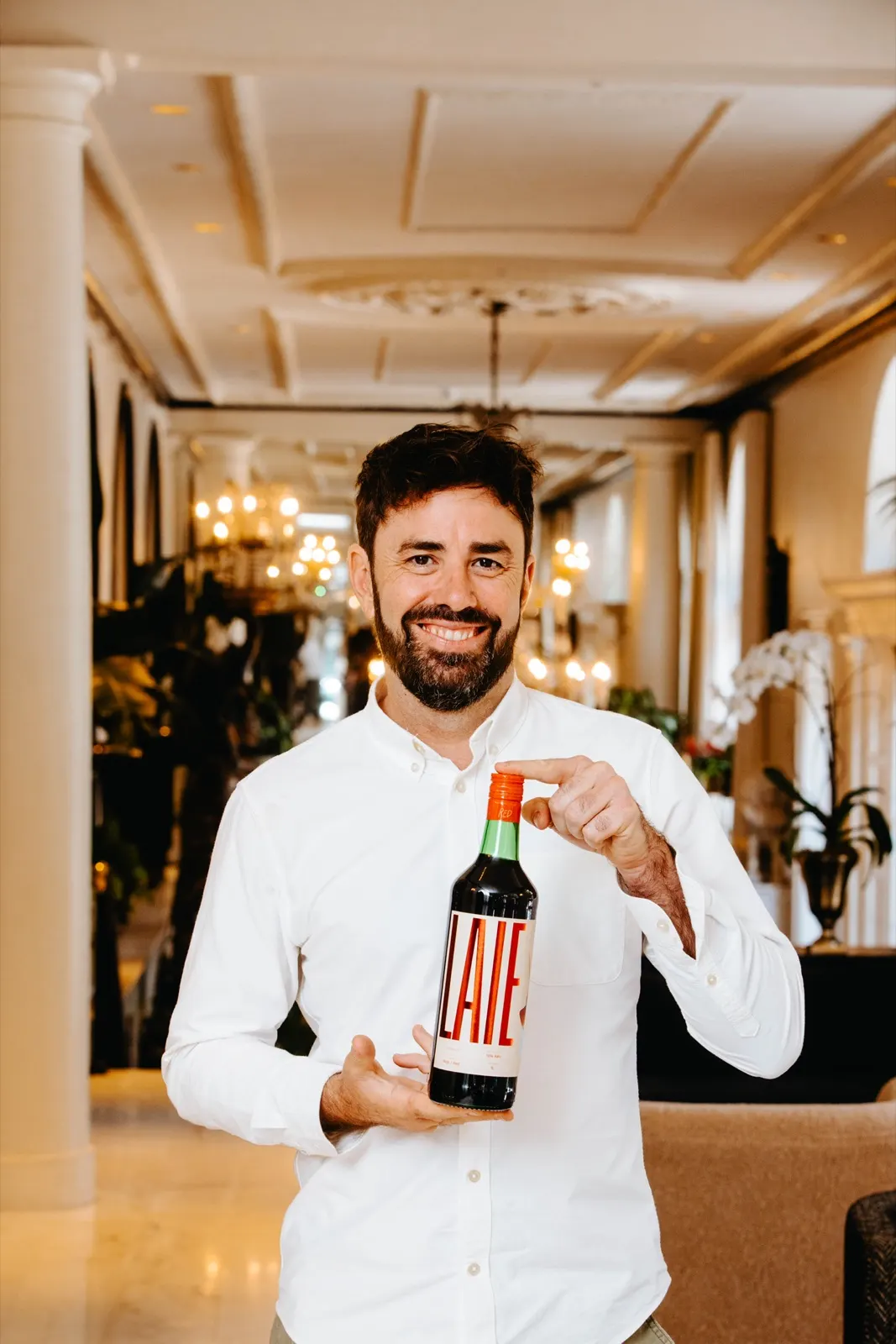 Àlex Cardona in a white shirt, smiling and holding a bottle of LAIE Rojo in a hotel lobby with chandeliers.