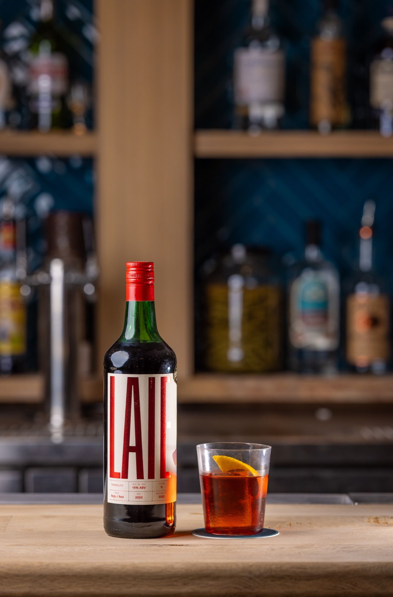 A LAIE Rojo Vermouth bottle beside a short glass of red-amber drink with an orange slice on a wooden bar, shelves blurred behind.
